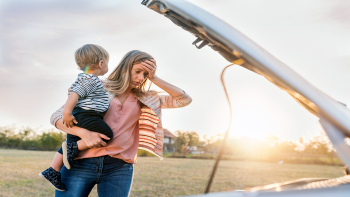 Portrait of young Caucasian woman standing by broken down car and waiting for assistance while holding her baby boy during sunset. Photo of female waits for assistance near her car broken down on the road side. Frustrated young brown hair woman looking at broken down car engine on street. Woman does not understand why her car engine failed. Desperate mother holding her angry baby.