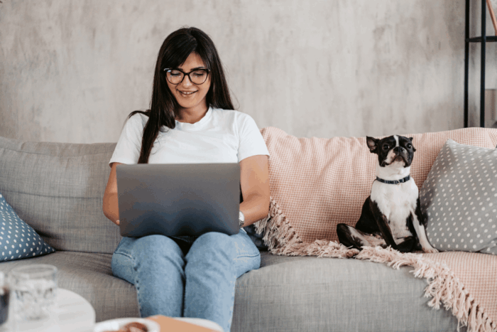 Young adult woman sitting on her couch with her laptop in her lap and dog sitting next to her