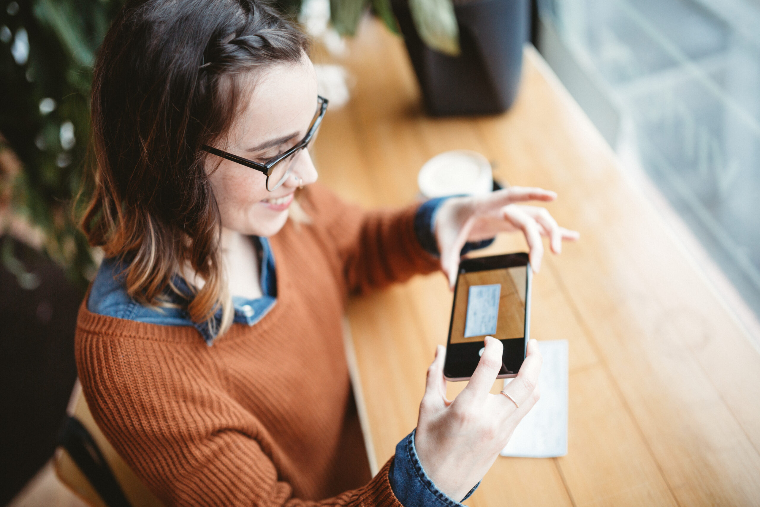 A smiling young woman takes a picture with her smart phone of a check or paycheck for digital electronic depositing, also known as "Remote Deposit Capture". She sits in a coffee shop, enjoying an espresso latte. Bright sunlight shines in the window. Horizontal image.